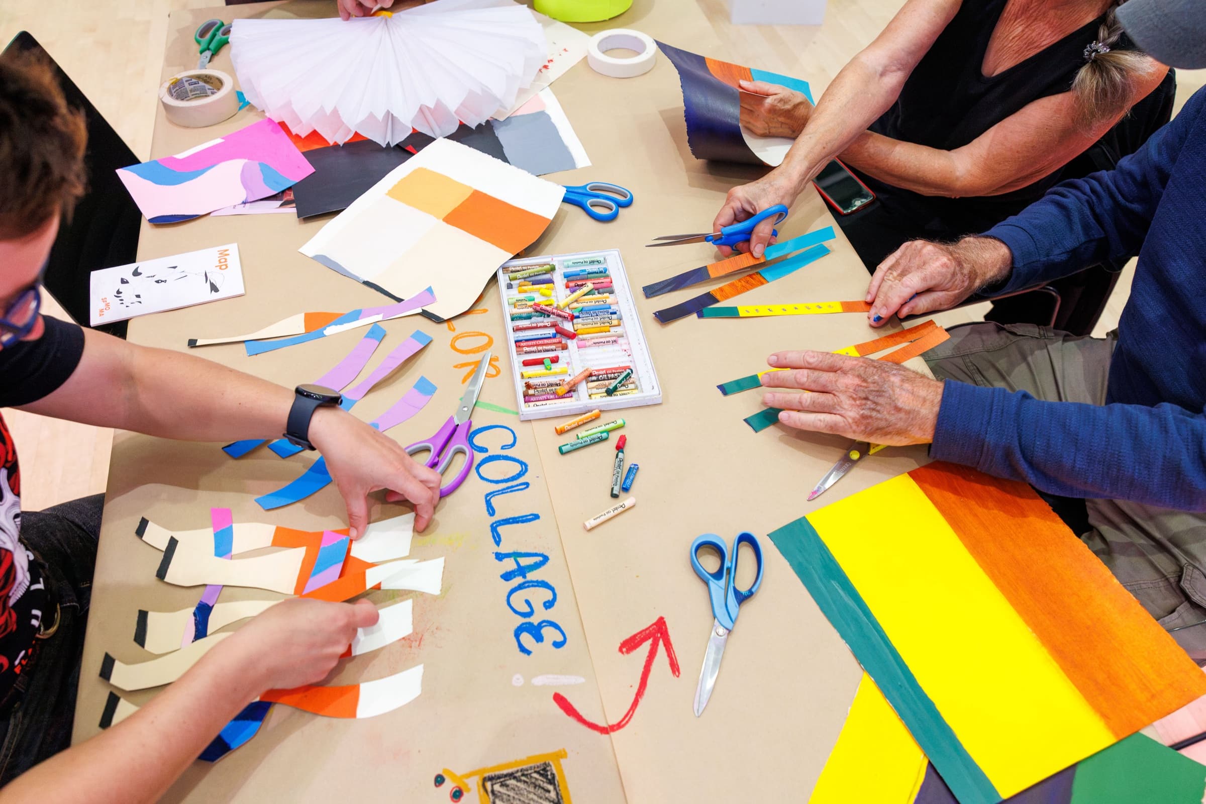 Overhead view of hands cutting colorful paper strips around a table with scissors, oil pastels, and the word “collage” written on it
