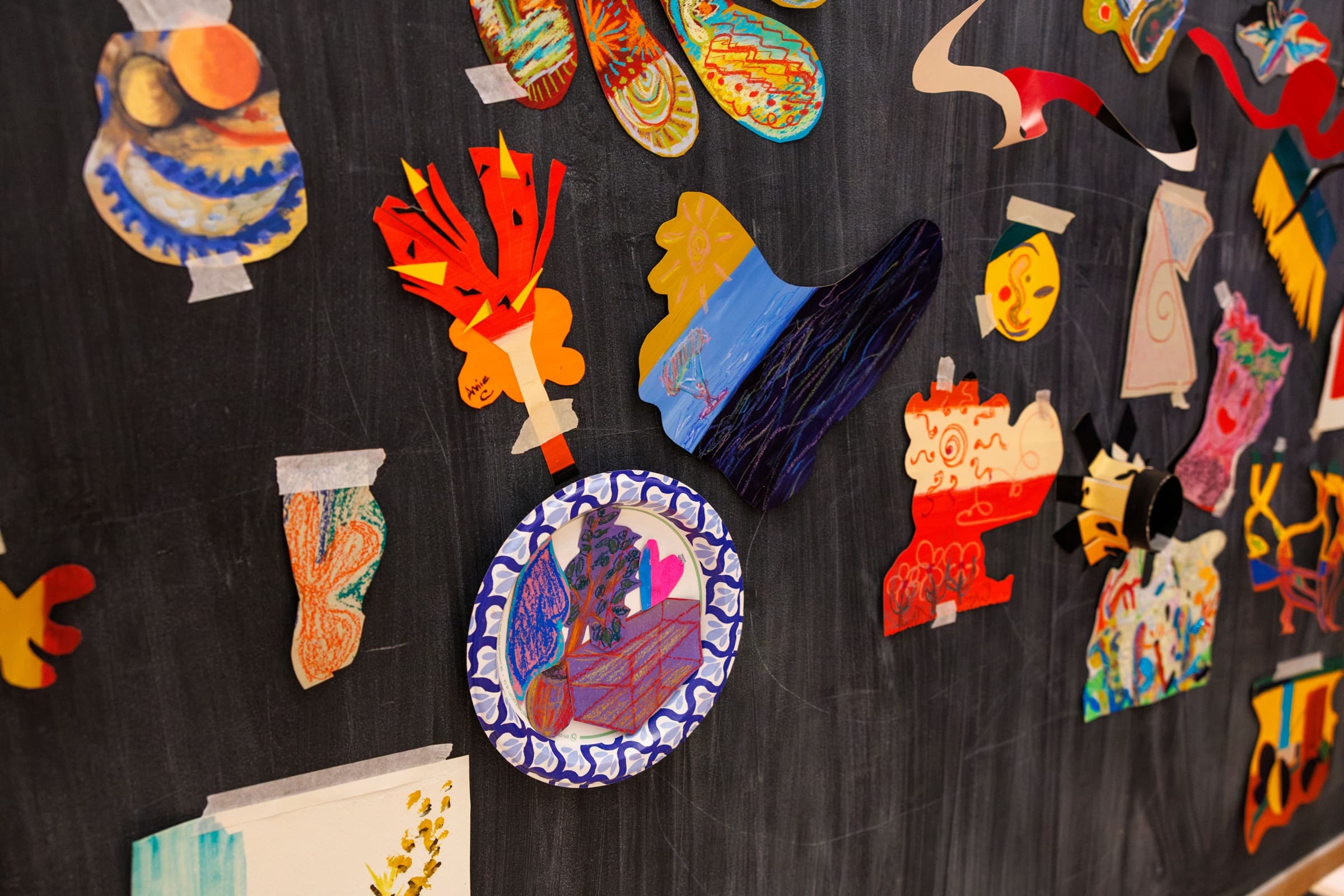 Close-up of colorful cut-paper shapes and pastel drawings taped to a dark wall, including one on a paper plate