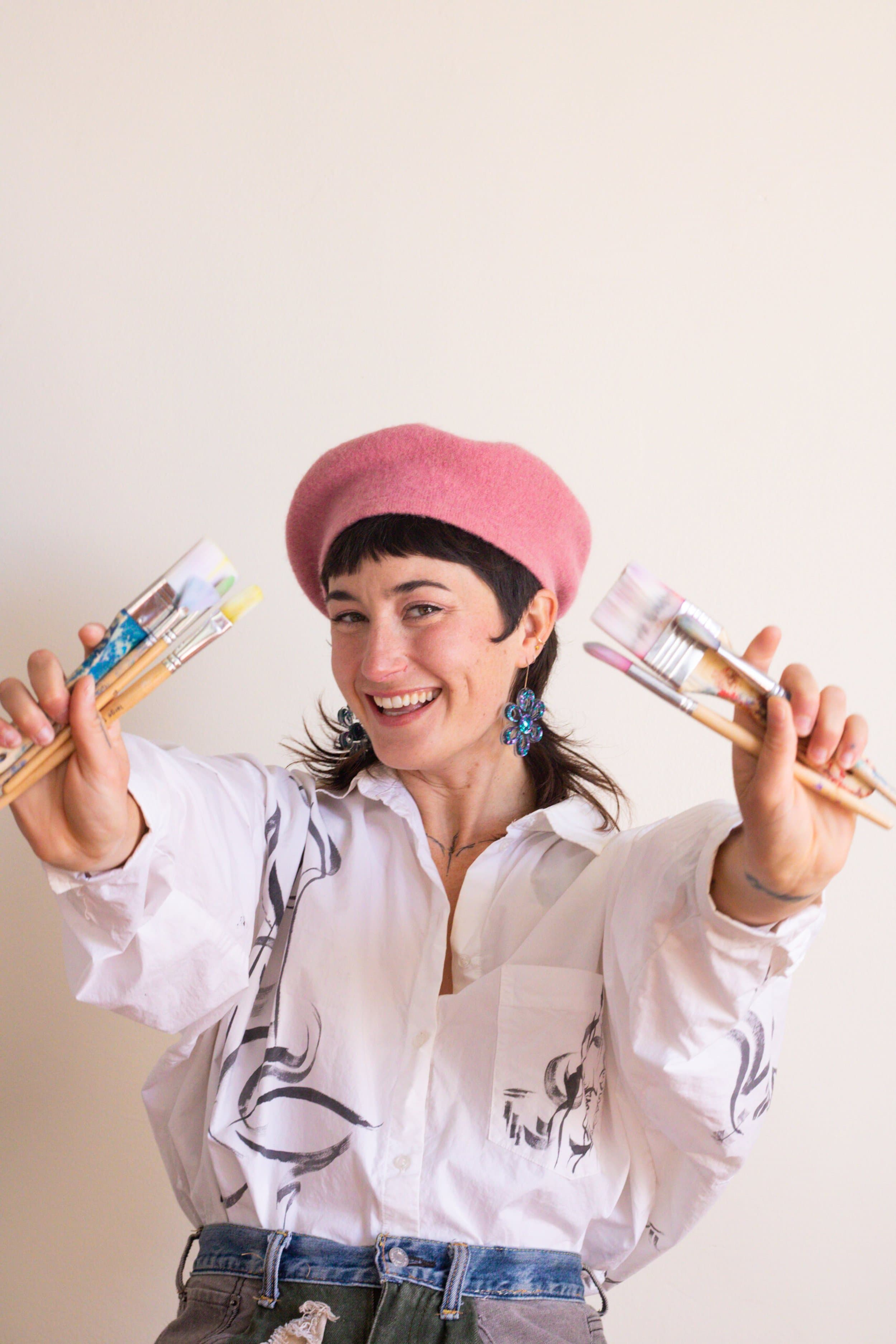 Jules wearing a pink beret and floral earrings, smiling while holding paintbrushes in both hands, dressed in a white shirt with black artistic designs.